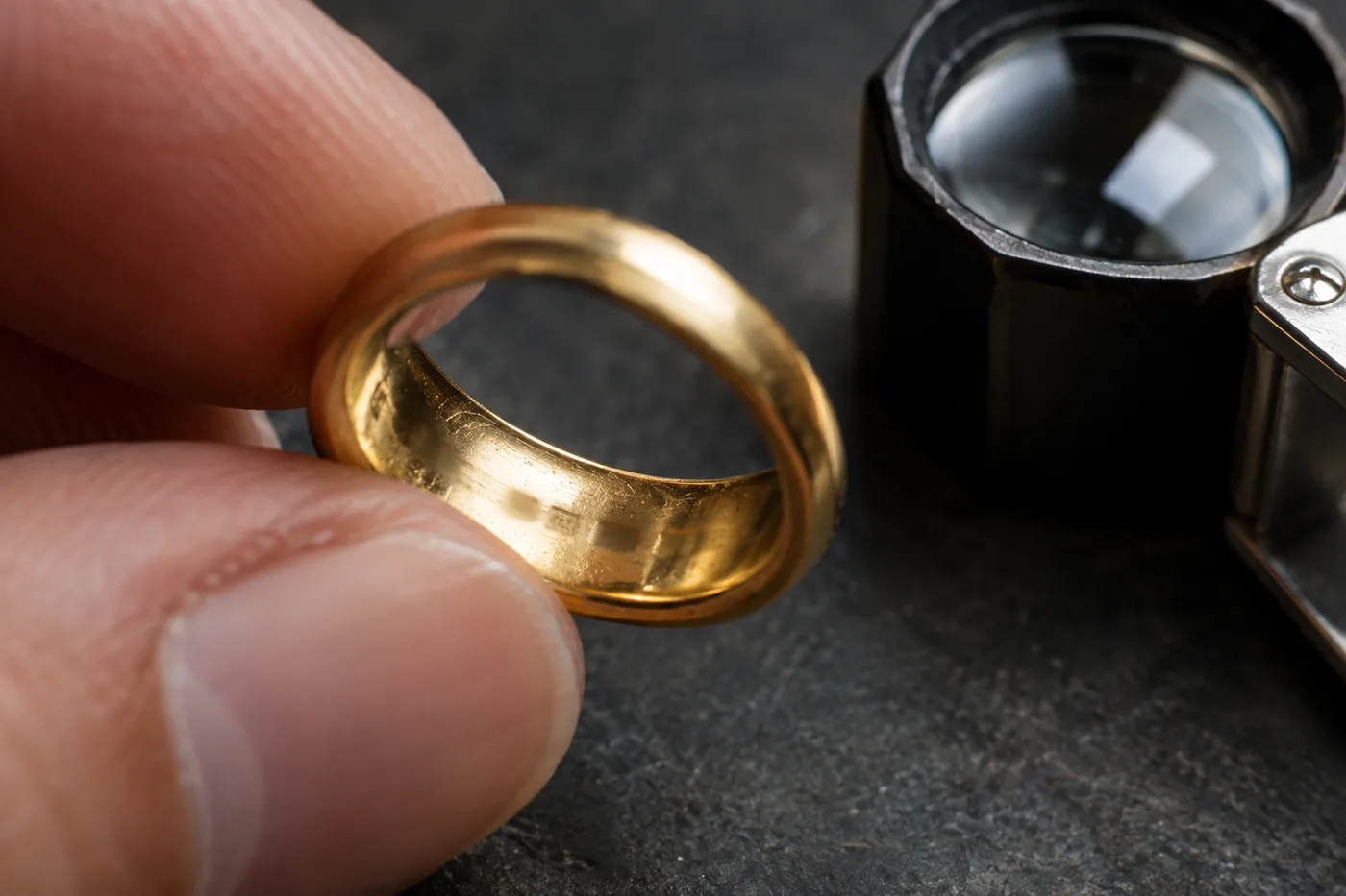 Bague en or tenue entre deux doigts, l'intérieur de l'anneau tourné vers une loupe de bijoutier